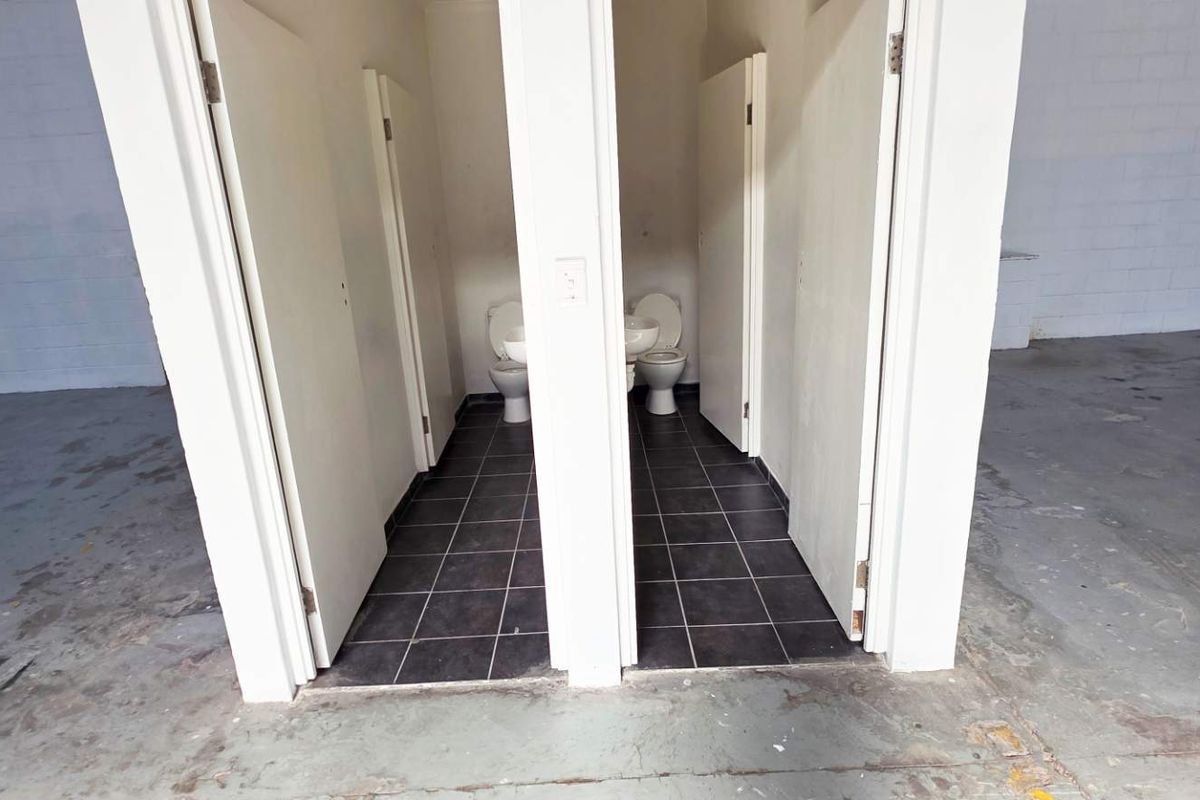 Two separate restroom cubicles featuring tiled flooring, situated within a large open-plan industrial or retail warehouse space with concrete flooring.