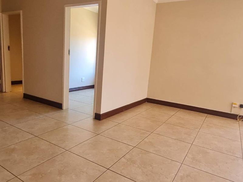 An empty living area featuring neutral ceramic floor tiles, coved ceiling cornices, and dark timber-look skirting boards, leading into an adjacent hallway.