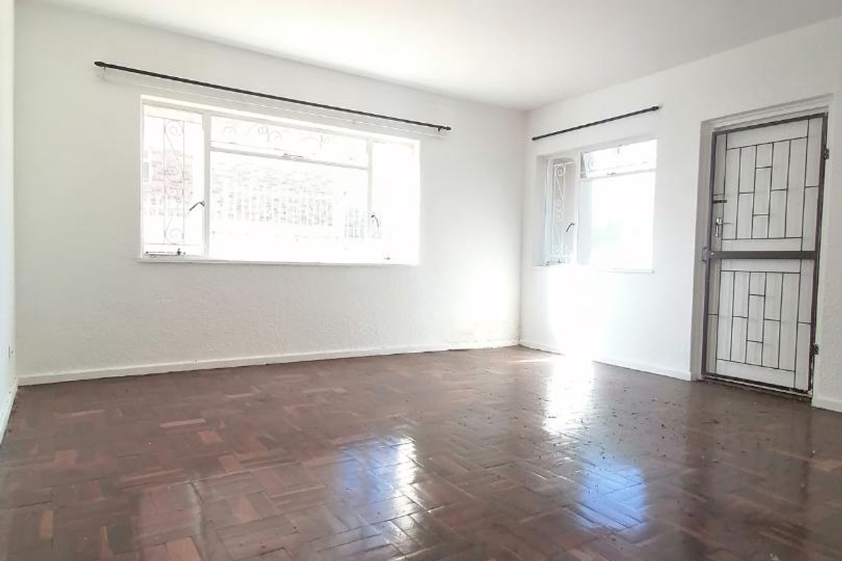 An empty living room featuring original parquet flooring, security gate, and large windows providing natural light.