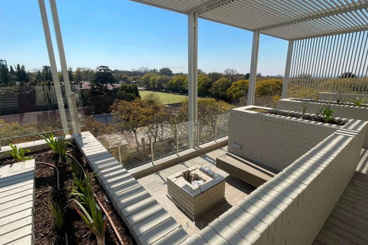 Spacious rooftop balcony featuring a modern louvred pergola, built-in brick planters, integrated fire pit, and glass balustrades with panoramic views.