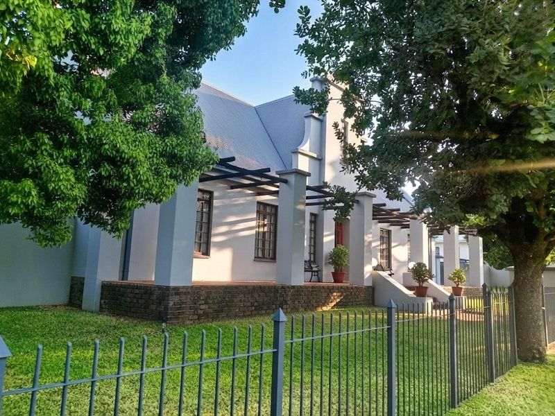 A charming Cape Dutch style home featuring a covered veranda, brick base, and a well-maintained garden with metal perimeter fencing.