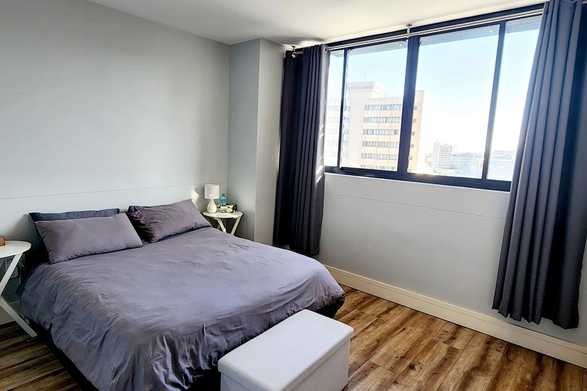 A bright bedroom featuring wood-look flooring, large windows with city views, and neutral wall finishes.