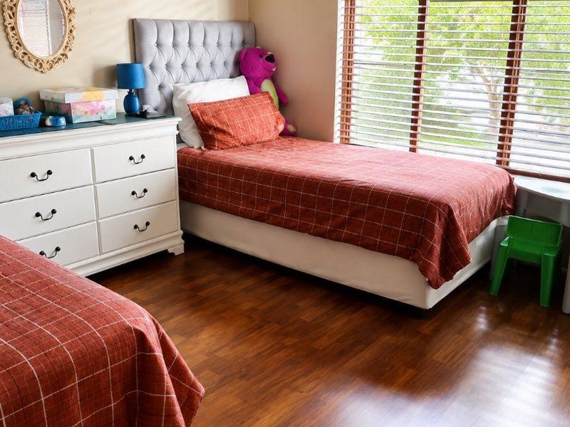 A well-lit bedroom featuring warm-toned laminate flooring and large windows fitted with venetian blinds.