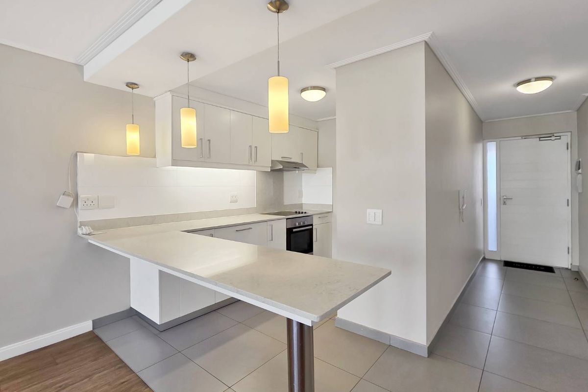 Modern open-plan kitchen featuring white cabinetry, stone countertops, a breakfast bar, and integrated appliances, seamlessly connected to the entrance hallway.