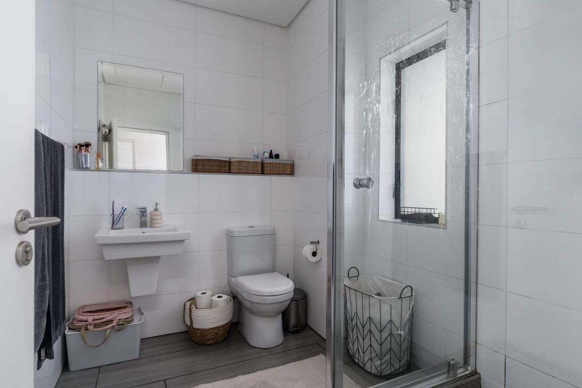 A clean, modern bathroom featuring white wall tiles, a wall-mounted basin, a toilet, and a glass-enclosed shower with a window.