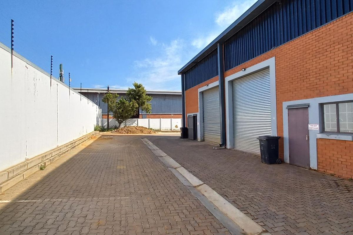 Exterior view of an industrial property featuring a paved yard, roller shutter doors, and perimeter wall with electric fencing.