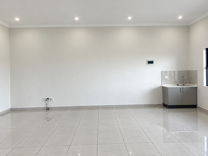 A bright, open-plan retail space featuring polished tile flooring, recessed ceiling lighting, and a compact kitchenette area with a sink and cabinetry.