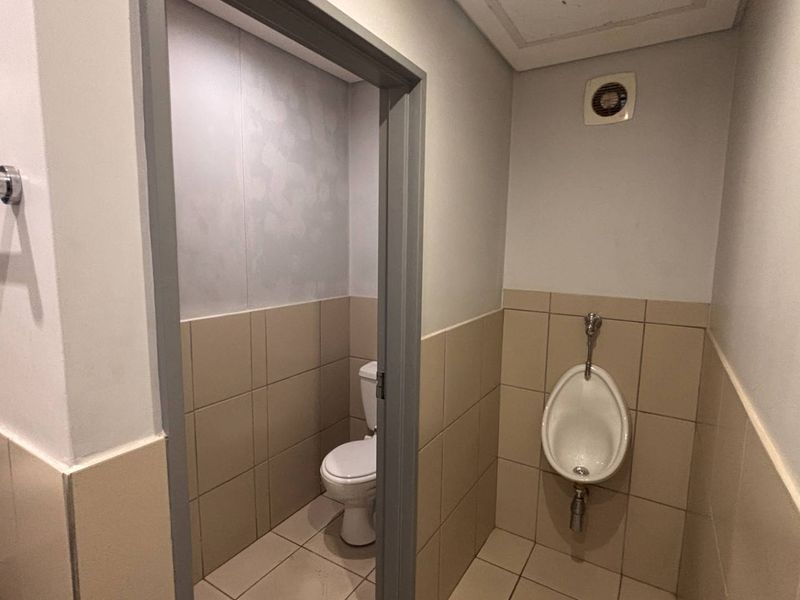 A functional office bathroom featuring a private toilet cubicle, wall-mounted urinal, tiled finishes, and an integrated extractor fan.