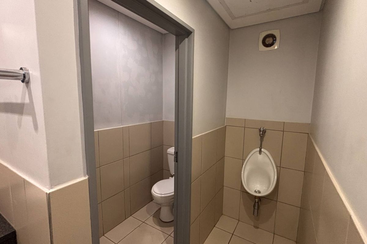 A functional office bathroom featuring a private toilet cubicle, wall-mounted urinal, tiled finishes, and an integrated extractor fan.