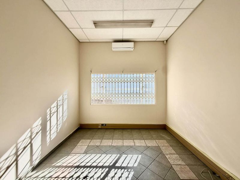 A vacant office space featuring a suspended ceiling, split-system air conditioning, security bars on the window, and tiled flooring.