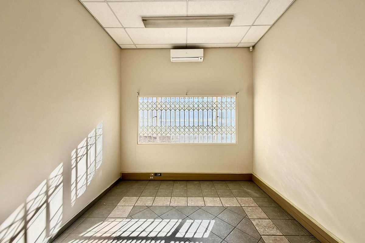 A vacant office space featuring a suspended ceiling, split-system air conditioning, security bars on the window, and tiled flooring.
