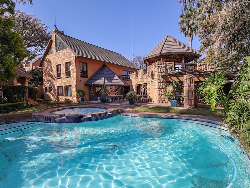 A spacious outdoor entertainment area featuring a large swimming pool with an integrated spa, stone-clad architecture, and a covered thatched gazebo.