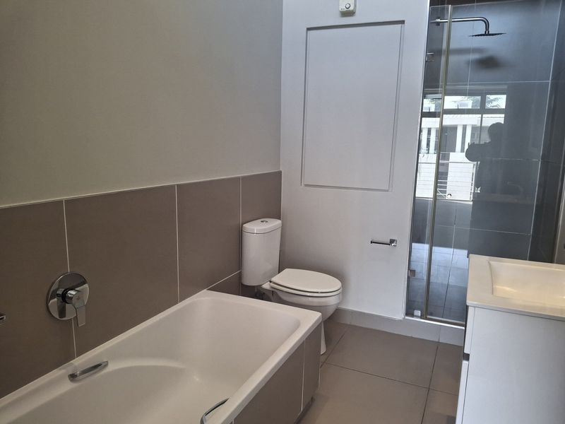 Modern bathroom featuring a bathtub, separate shower enclosure with rain shower head, toilet, and vanity basin with neutral wall and floor tiling.