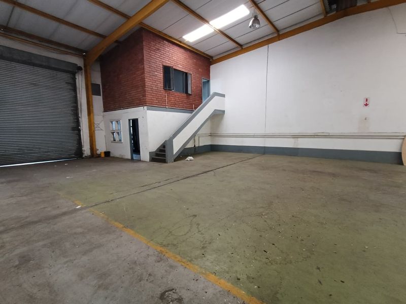 Spacious industrial warehouse unit featuring a high-clearance roller shutter door, concrete flooring, and an integrated mezzanine level with staircase access.