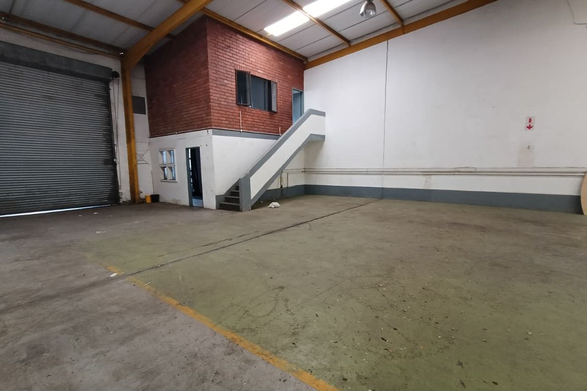 Spacious industrial warehouse unit featuring a high-clearance roller shutter door, concrete flooring, and an integrated mezzanine level with staircase access.