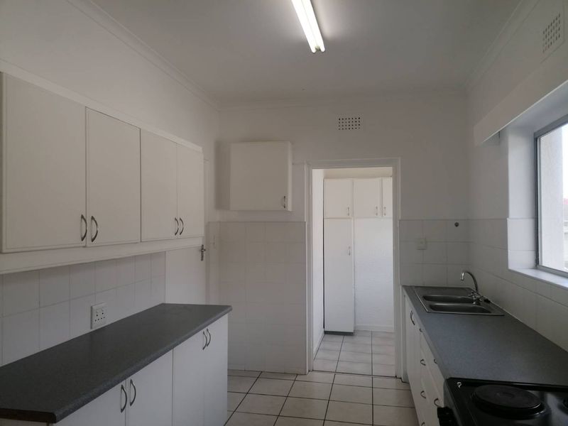 A functional, empty kitchen featuring white cabinetry, dark laminate countertops, tiled flooring, and a double bowl sink with an adjacent scullery area.