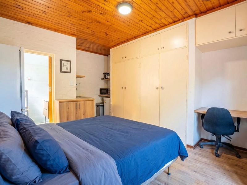 A bedroom featuring built-in cupboards, overhead storage, a built-in desk, and a timber ceiling, with an adjacent kitchenette area.