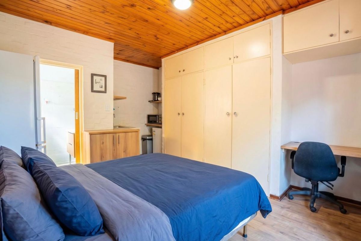 A bedroom featuring built-in cupboards, overhead storage, a built-in desk, and a timber ceiling, with an adjacent kitchenette area.