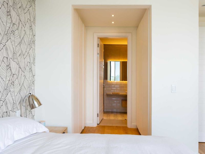 A modern bedroom featuring hardwood flooring, built-in cupboards, and a clear view into an adjacent en-suite bathroom with contemporary finishes.