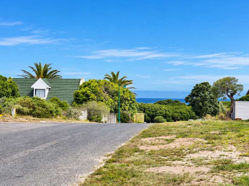 A street-level view of a residential vacant plot featuring a paved road, boundary wall, and distant ocean views.