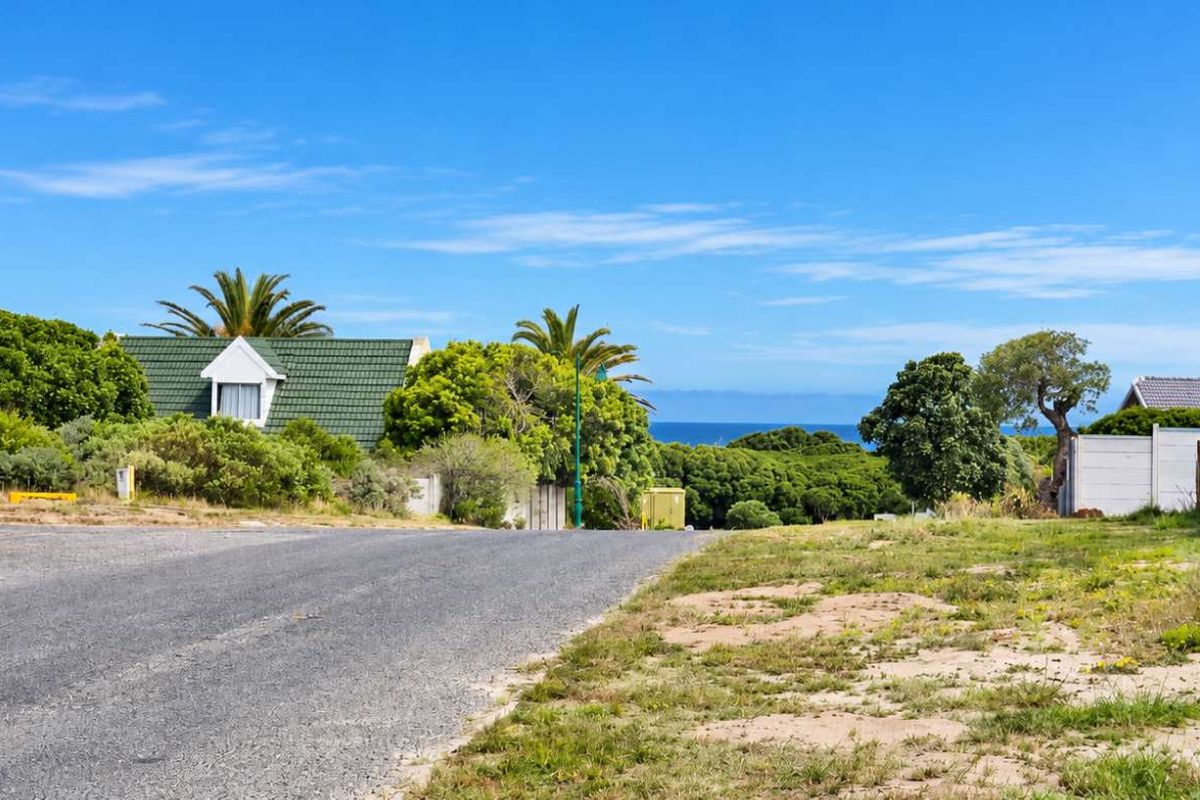 A street-level view of a residential vacant plot featuring a paved road, boundary wall, and distant ocean views.