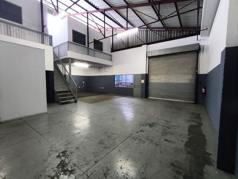 Spacious industrial warehouse unit featuring a high-clearance ceiling with exposed trusses, a mezzanine level, concrete flooring, and a large roller shutter door.