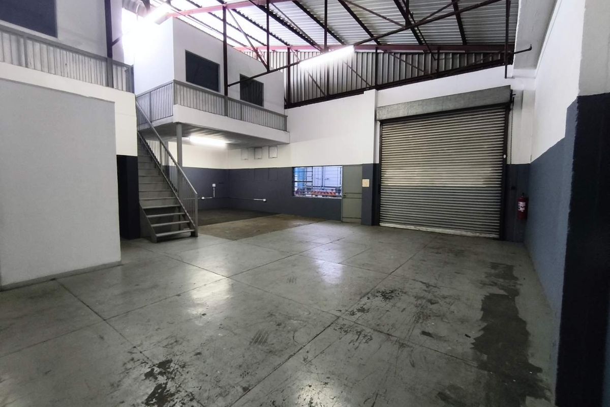 Spacious industrial warehouse unit featuring a high-clearance ceiling with exposed trusses, a mezzanine level, concrete flooring, and a large roller shutter door.