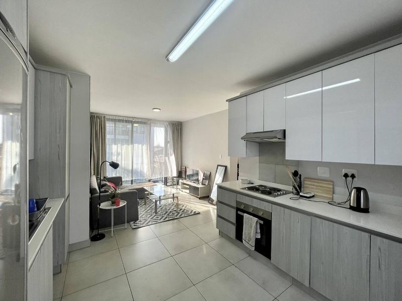 Modern open-plan living area featuring a contemporary kitchen with built-in appliances and tiled flooring, seamlessly flowing into a bright lounge with balcony access.