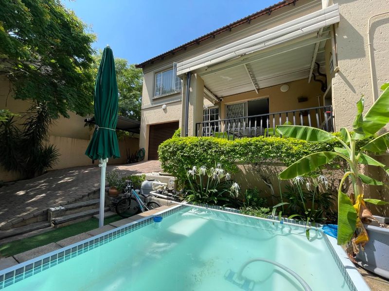 Rear exterior view of the townhouse featuring a swimming pool, covered patio area, and paved driveway with established garden landscaping.