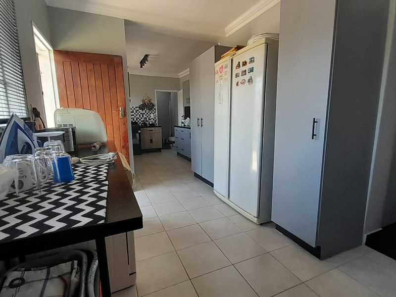 A functional kitchen area featuring built-in cabinetry, tiled flooring, and natural light through venetian blinds, leading into an adjacent scullery or utility space.