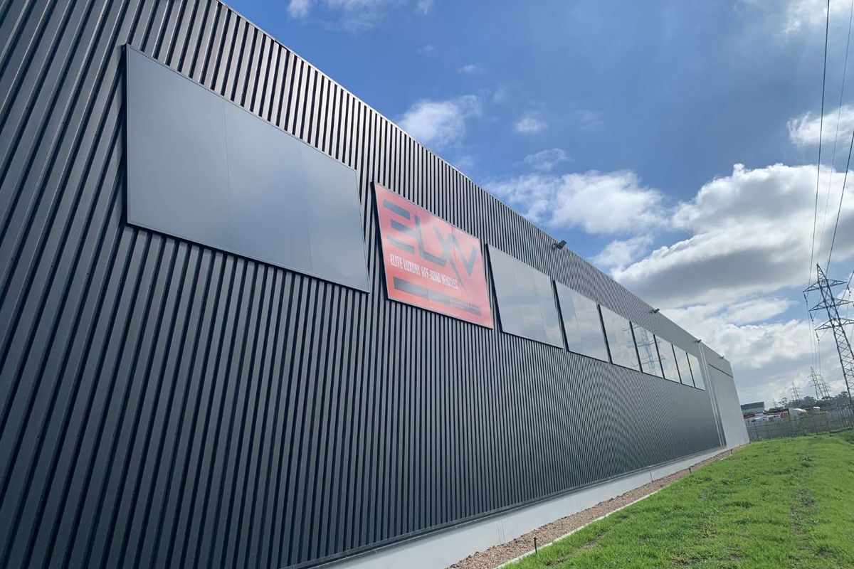 Exterior view of a modern industrial building featuring dark corrugated metal cladding, large windows, and prominent signage.