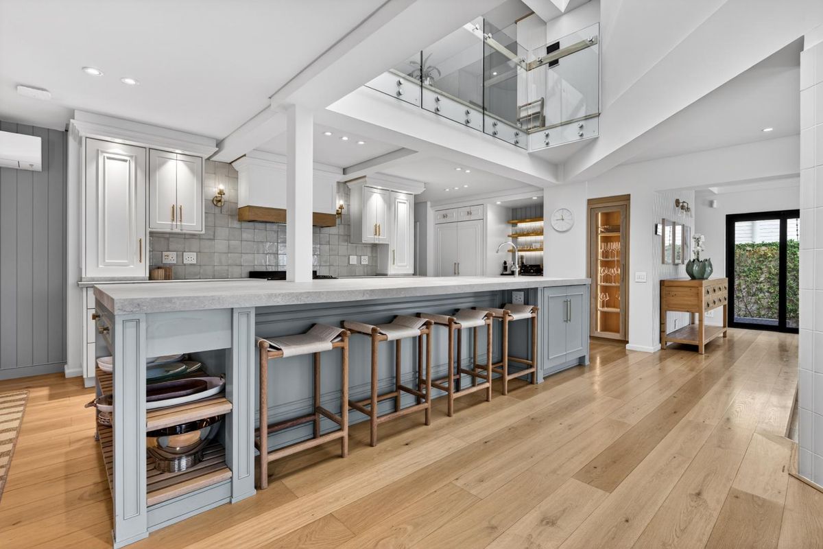 Modern, high-end kitchen featuring a large island with stone countertops, shaker cabinetry, hardwood floors, and a striking glass-enclosed mezzanine level above.