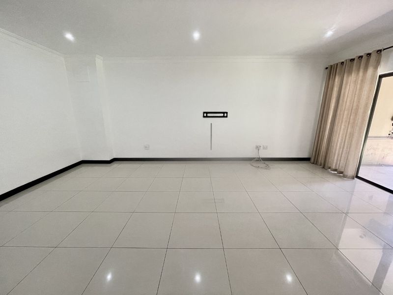 A bright, empty living room featuring neutral tiled flooring, recessed downlighting, and a wall-mounted TV bracket, leading to an outdoor area via sliding doors.