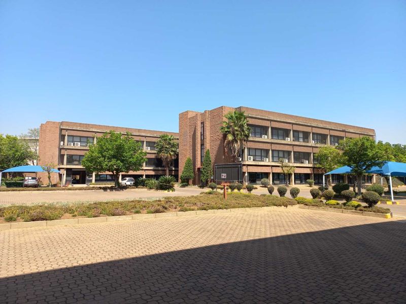 A multi-story brick office building featuring a paved parking area, landscaped grounds, and shaded parking bays under a clear blue sky.