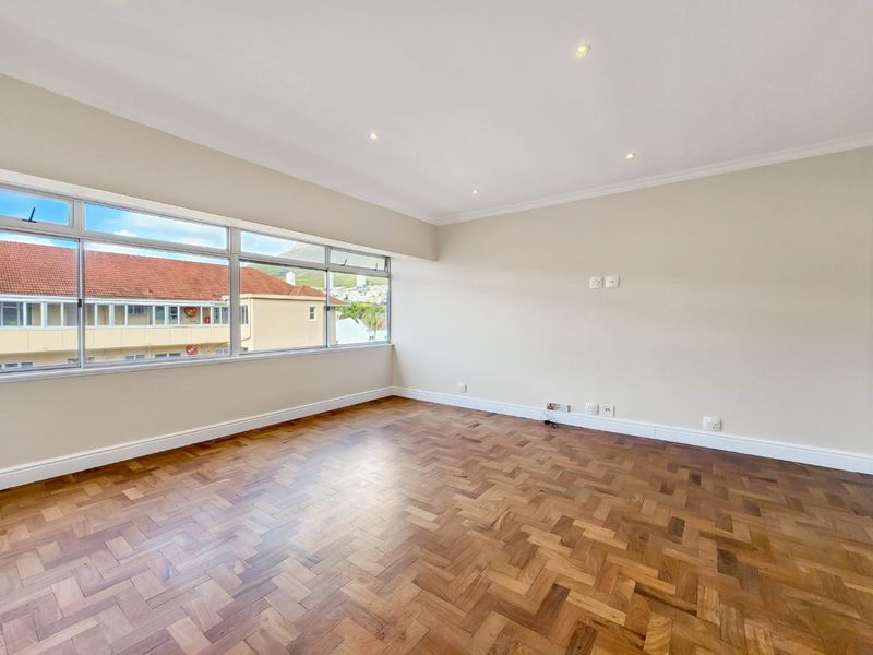 A bright and spacious living area featuring classic herringbone parquet flooring, recessed lighting, and large windows offering natural light and neighborhood views.