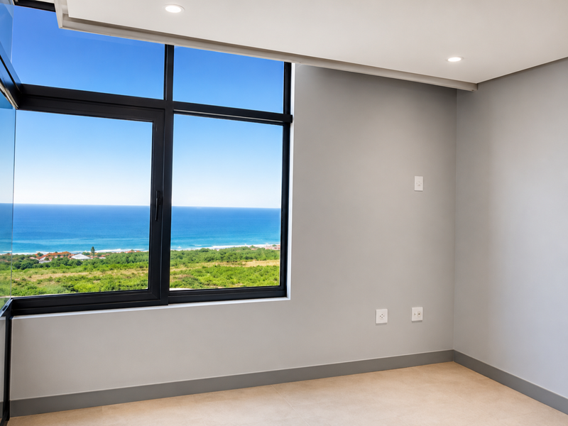 A bright, empty bedroom featuring large aluminium-framed windows with expansive ocean and coastal views, neutral wall finishes, and modern tiled flooring.