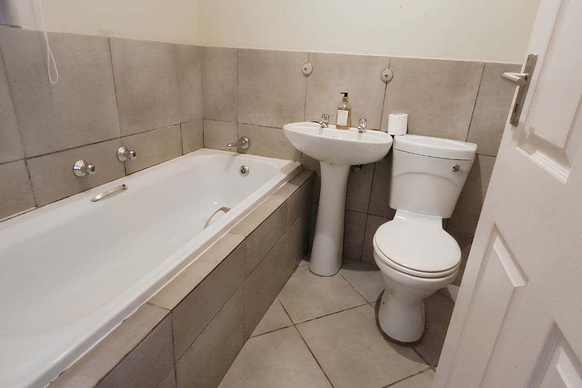A functional bathroom featuring a built-in bathtub, pedestal basin, and toilet with neutral ceramic wall and floor tiling.
