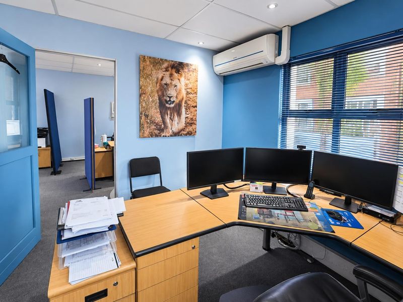 A functional office space featuring carpeted flooring, a suspended ceiling with recessed lighting, an integrated air conditioning unit, and large windows with venetian blinds.