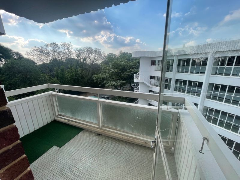A balcony area featuring tiled flooring, a white railing, and a glass balustrade with a visible crack, overlooking the surrounding apartment complex and trees.