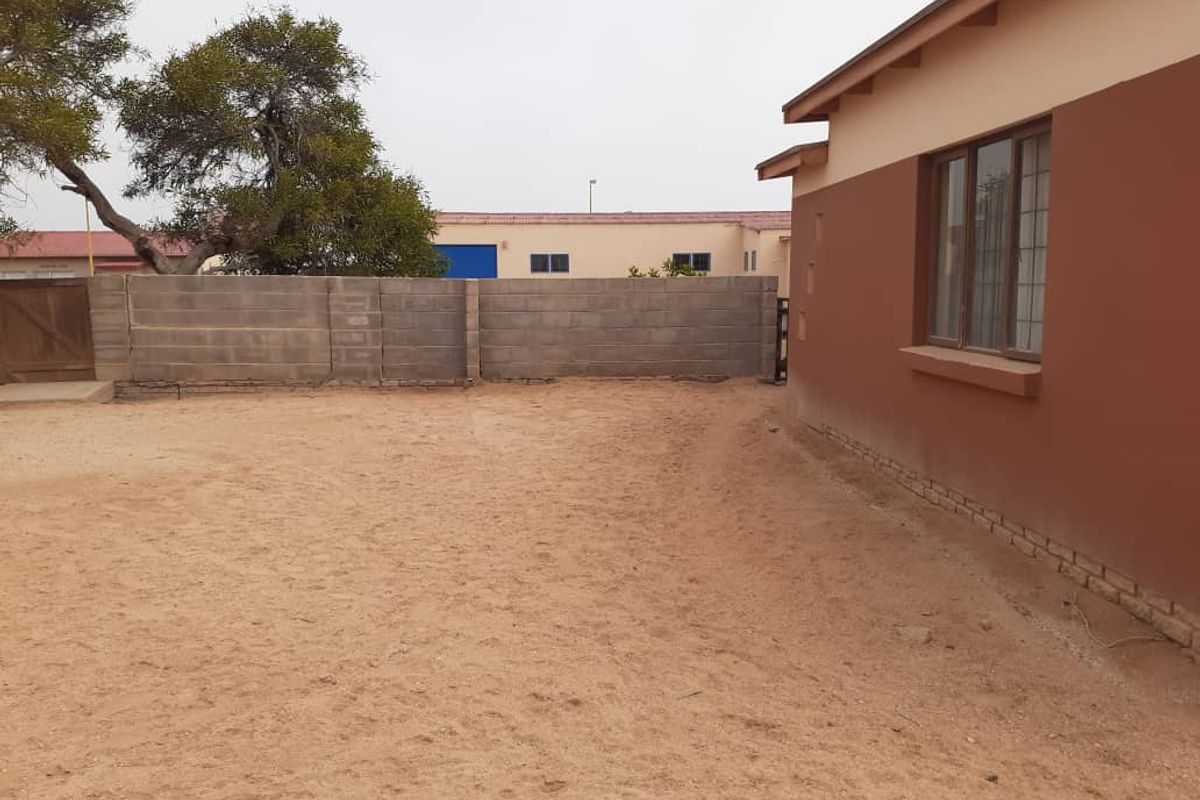 Exterior view of the property showing a side facade with window and an expansive, unpaved sandy yard area enclosed by a concrete block wall.