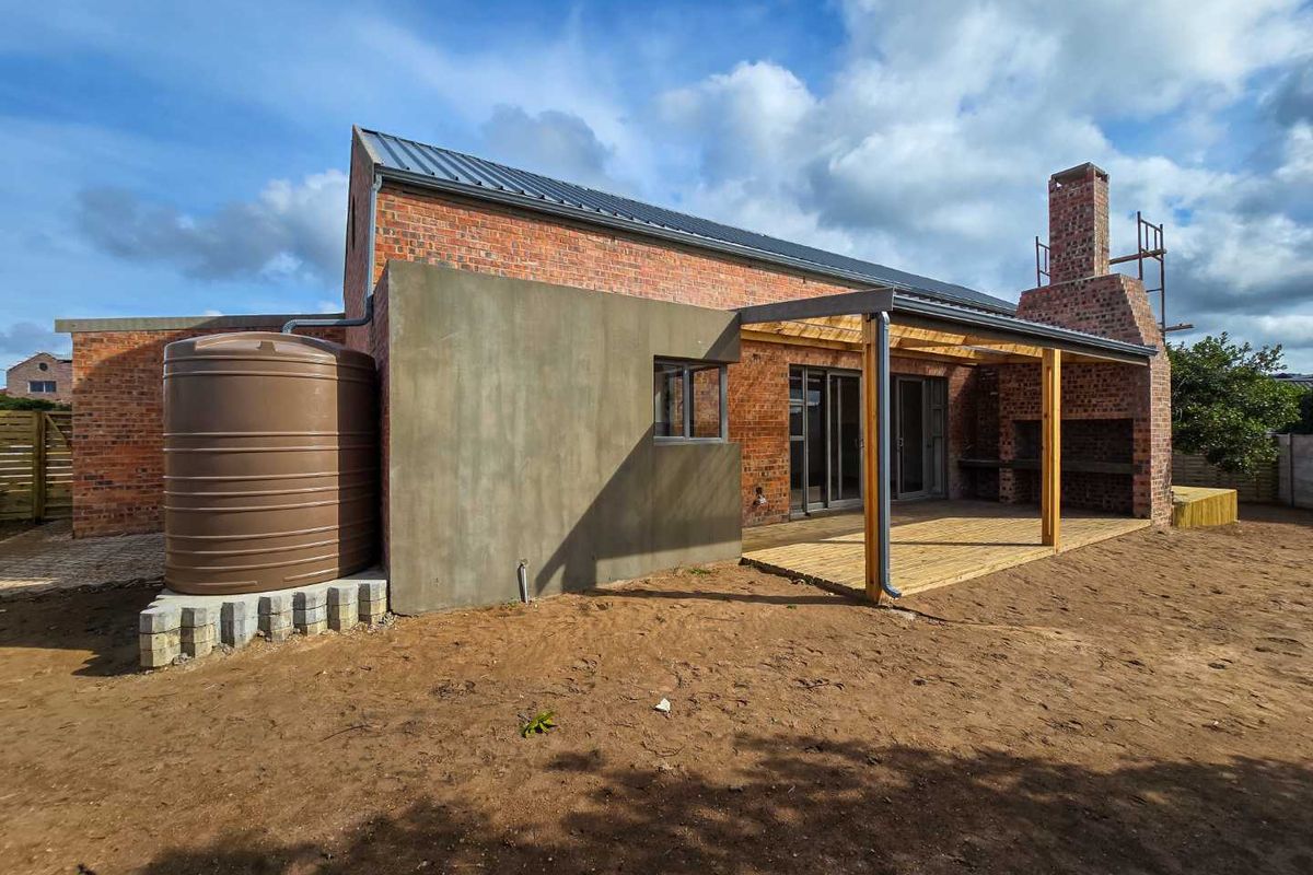 Rear view of a modern brick townhouse featuring a covered timber deck, built-in braai, and a large water storage tank in an unfinished garden.