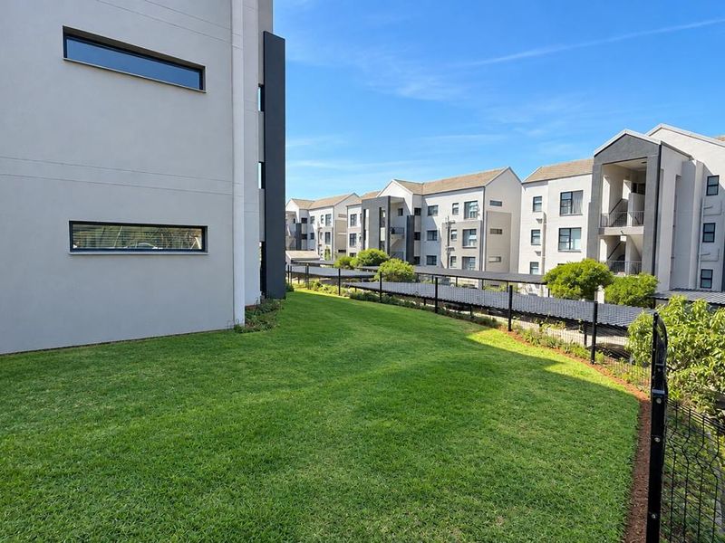 A well-maintained communal garden area featuring manicured lawns, modern apartment building facades, and covered parking bays within a secure residential complex.
