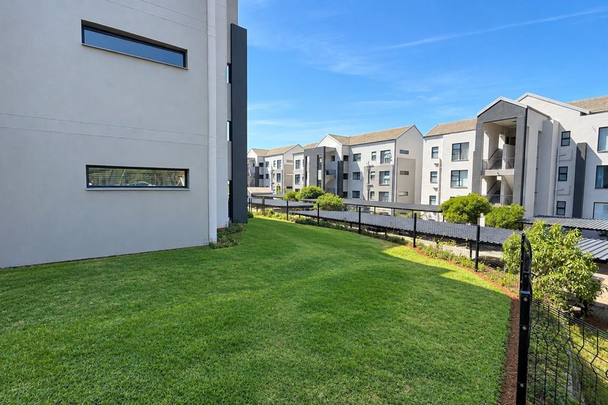 A well-maintained communal garden area featuring manicured lawns, modern apartment building facades, and covered parking bays within a secure residential complex.