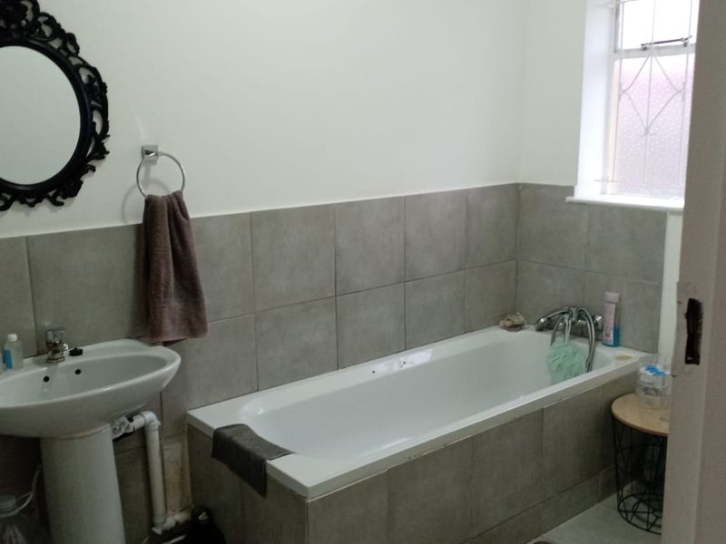 A functional bathroom featuring a tiled bathtub, pedestal basin, and neutral wall tiling with an obscured glass window for privacy.