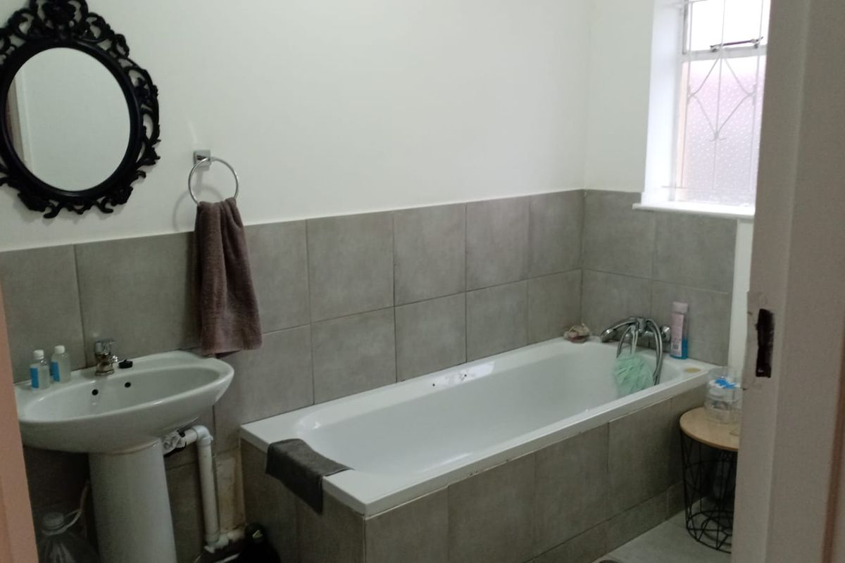 A functional bathroom featuring a tiled bathtub, pedestal basin, and neutral wall tiling with an obscured glass window for privacy.