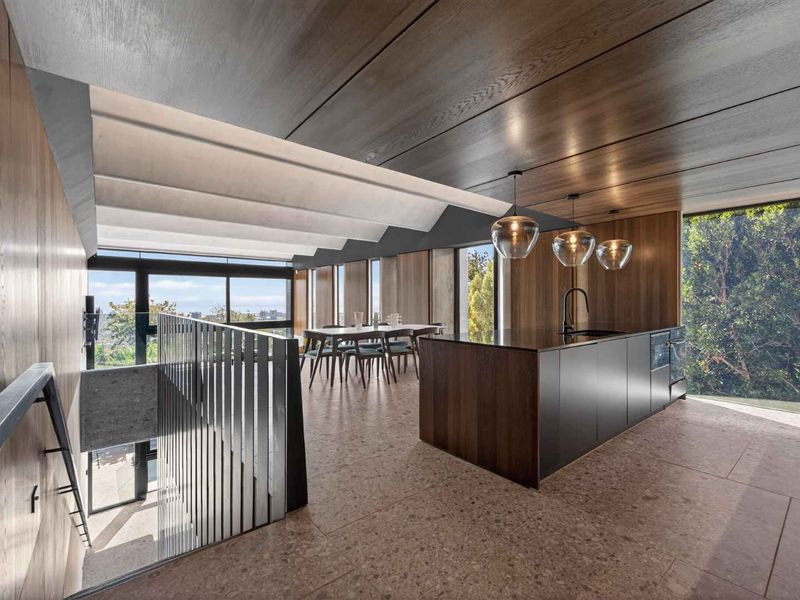 Modern open-plan living area featuring a sleek kitchen island, timber-paneled ceilings, terrazzo flooring, and expansive windows overlooking lush greenery.