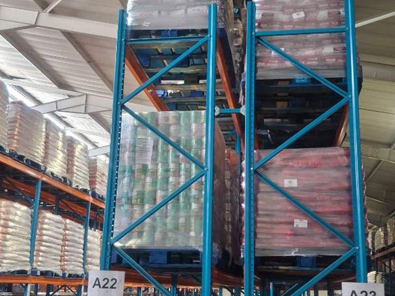 Industrial warehouse interior featuring high-level steel pallet racking systems for bulk storage and exposed structural roof trusses.