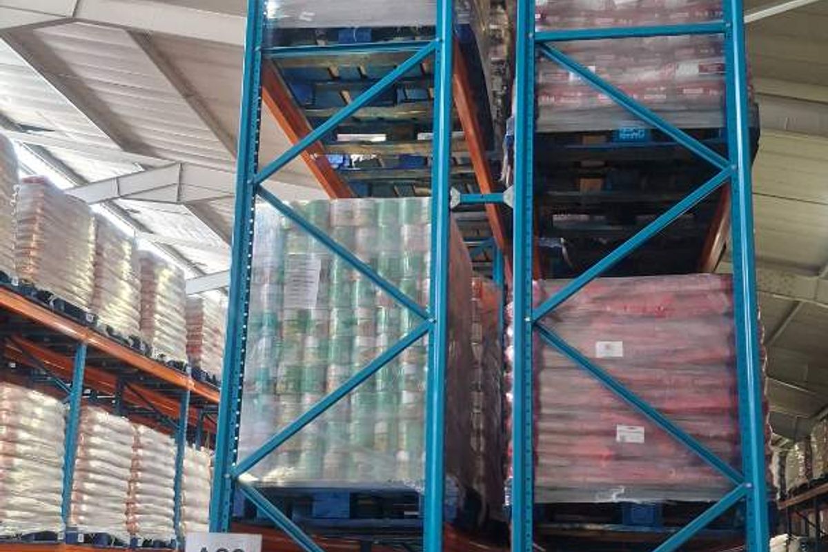 Industrial warehouse interior featuring high-level steel pallet racking systems for bulk storage and exposed structural roof trusses.