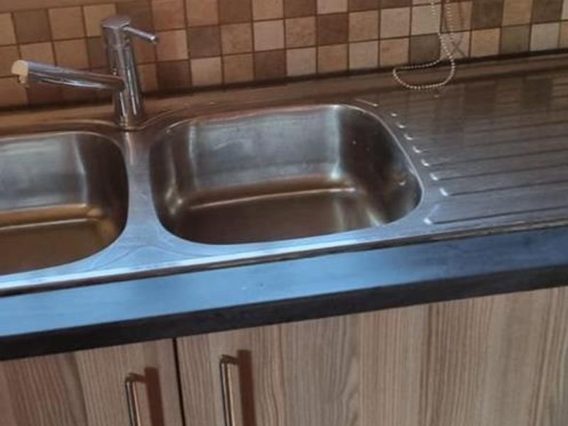A functional kitchen area featuring a double bowl stainless steel sink, mosaic tile backsplash, and modern laminate cabinetry with a dark countertop.