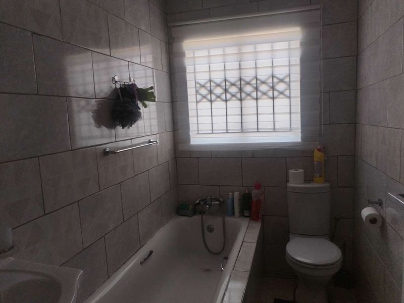 A functional bathroom featuring a built-in bathtub, toilet, and full-height wall tiling, with a window fitted with a security gate.